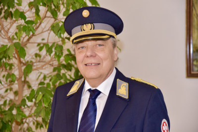 Ein Mann in Uniform mit Hut lächelt vor einem Pflanzenhintergrund.